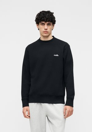 Νεαρός άνδρας με σγουρά σκούρα μαλλιά που φοράει μαύρο φούτερ μακρυμάνικο με μικρό λευκό λογότυπο και ανοιχτό γκρι παντελόνι φόρμας, ουδέτερο φόντο.