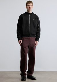 Hanorac negru cu fermoar, guler cu revere și două buzunare frontale, asortat cu pantaloni bordo cu logo subtil. Întregul outfit este completat de pantofi negri.