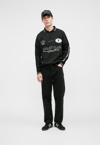 Črna polo majica z dolgimi rokavi z belimi poudarki in besedilom, kombinirana s črnimi kavbojkami in črnimi supergami z belimi črtami.