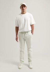 T-shirt en coton blanc, coupe ample ; pantalon léger vert avec cordon de serrage ; baskets blanches ; casquette beige avec logo ; texture lisse, style décontracté.