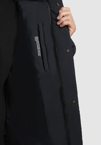 Tumes sinine polsterdatud jope, millel on tõmblukuga sulgemine, peidetud sisetasku ja silt hooldusjuhistega. Sile tekstuur.