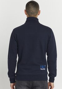 Tamno plavi dukserica s visokim ovratnikom, rebrastim manžetama i donjim rubom, koja na donjem dijelu leđa ima plavi izvezeni logo. Tkanina je mješavina pamuka.