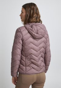 Veste matelassée violette en forme de doudoune avec un motif en chevron, capuche réglable, et un écusson de logo sur la manche. Texture lisse et design ajusté.