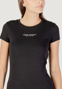 T-shirt nera a maniche corte in cotone, con scollatura rotonda e testo stampato bianco "ARMANI EXCHANGE MILANO/NEW YORK" sul petto.