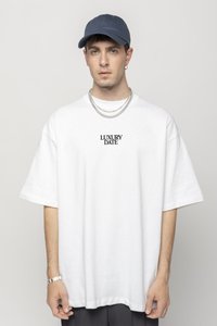 Camiseta blanca de gran tamaño con el texto negro "LUXURY DATE", combinada con una gorra azul y cadenas de plata en capas. Fondo sencillo.