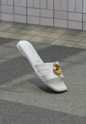 Biela šľapka s krokodílovou textúrou, zlatým kruhovým ozdobením, štvorcovou špičkou a logom "ALDO" na vložke, na pozadí sivej dlažby.