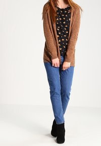 Brązowy dziany kardigan noszony na czarnej bluzce w kwiatowy wzór, zestawiony z niebieskimi dżinsami i czarnymi botkami za kostkę. Kardigan jest otwarty z przodu.