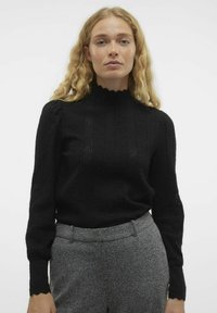 Zwarte gebreide trui met hoge scalloped hals, geavanceerd patroon en lange mouwen. Gepaard met grijze getextureerde broek.