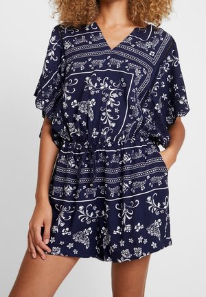 Kobieta w granatowym kombinezonie w kwiaty, z dekoltem w kształcie litery V, wcięciem w talii i krótkimi, luźnymi rękawami, z jedną ręką w kieszeni.