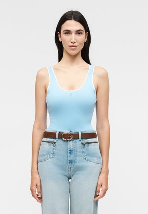 Femme aux longs cheveux foncés portant un top sans manches bleu clair, une ceinture marron et un jean taille haute bleu clair, debout devant un fond uni.