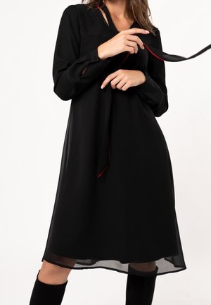 Vrouw draagt een zwarte midi-jurk met lange mouwen en houdt de dunne ceintuur met rode bies vast tegen een witte achtergrond.