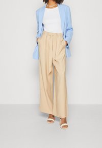 Lichtblauw colbert over een witte top, gecombineerd met beige, high-waisted, wijd uitlopende broek. Witte open-toe schoenen maken de outfit compleet.