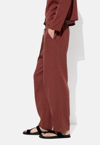 Rdzawoczerwone bawełniane spodnie o luźnym kroju, z bocznymi kieszeniami i elastycznym paskiem w talii, w połączeniu z czarnymi sandałami na paski.