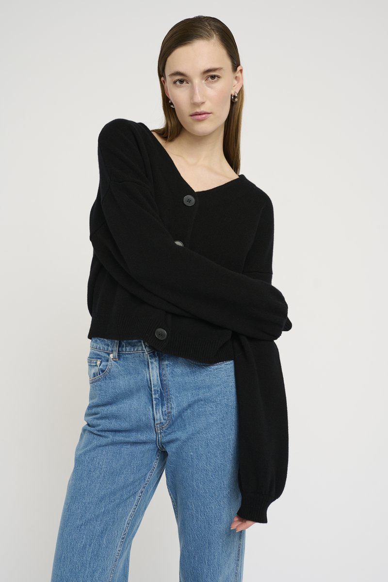 Cardigã preto em malha com decote em V, apresentando dois botões e mangas oversized, combinado com jeans de denim azul claro. Textura suave.