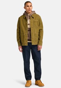 Olijfgroene jack met zakken, bruine hoodie eronder, marineblauwe cargobroek en gele laarzen. Eenvoudig ontwerp met knopen en subtiele stiksels.