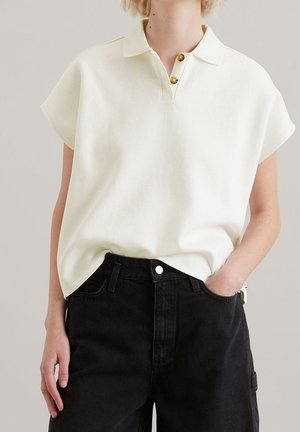 Personne portant une chemise blanche à manches courtes avec col et deux boutons, ainsi qu'un jean noir taille haute, une main dans la poche.