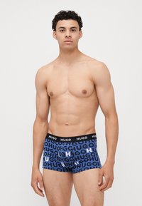 Blauwe boxershorts met zwarte tailleband, voorzien van herhaaldelijk "HUGO" logo in zwart en blauw. Rekbare stof, aansluitend ontwerp. Model staat rechtop.