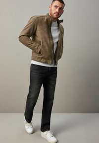 Chaqueta de cuero marrón con cuello alzado, cierre de cremallera y bolsillos laterales, combinada con un suéter gris, jeans negros y zapatillas blancas.