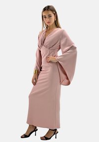 Vestido largo rosa con un escote en V profundo y mangas acampanadas, confeccionado con una tela suave. Combinado con sandalias de tacón negro.