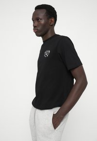 Μαύρο βαμβακερό t-shirt με μικρή λευκή γραφιστική σχεδίαση στο αριστερό στήθος, κοντά μανίκια και στρογγυλή λαιμόκοψη. Το μοντέλο φορά γκρι φόρμες.