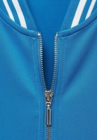 Giacca blu con zip e colletto a costine, caratterizzata da strisce bianche. La zip è metallica e ha un comodo tirazip. Tessuto liscio e strutturato.