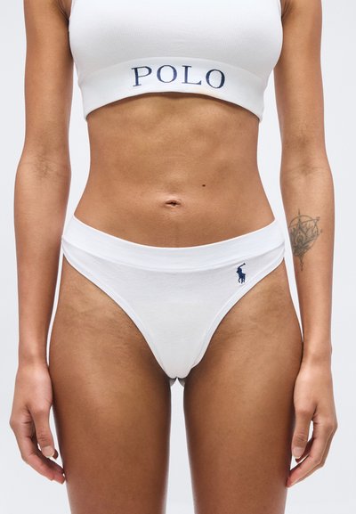 Femme portant un ensemble de sous-vêtements blanc de la marque Polo, montrant le ventre et les bras avec un tatouage détaillé sur l'avant-bras droit.