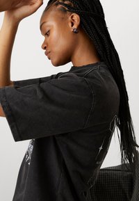 Black cotton t-shirt with short sleeves, distressed edges, and a graphic design on the side. Styled with braided hair and stud earrings.