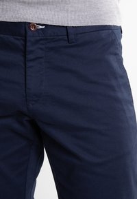 Námořnické chinos vyrobené z lehkého materiálu mají plochý přední díl, hnědý knoflík a kontrastní šití. Jednoduchý a univerzální design.