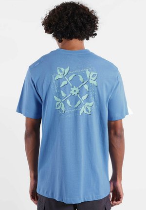 Homme aux cheveux bouclés portant un T-shirt bleu avec un motif floral symétrique vert dans le dos, debout devant un fond blanc.