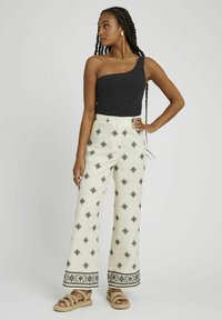 Haut noir asymétrique en tissu texturé, associé à un pantalon large crème avec des broderies géométriques noires et un ourlet à motifs. Bretelles tressées.