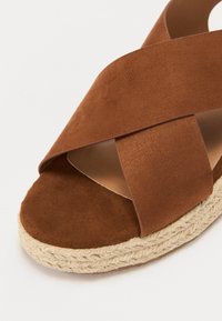 Brown suede espadrille sandal with crossed straps and a jute rope sole. Soft texture, open-toe design, and cushioned footbed.