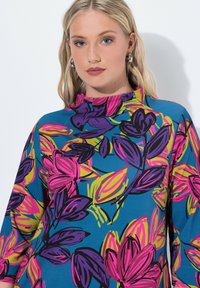 Wielokolorowa bluzka wykonana z niebieskiego materiału, ozdobiona wyraźnymi różowymi, purpurowymi i zielonymi wzorami kwiatowymi. Wysoki kołnierz i rękawy do trzech czwartych dodają szczegółów.