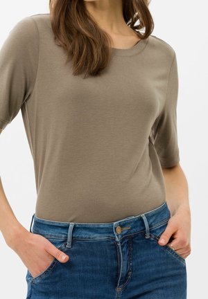 Woman wearing short-sleeve taupe t-shirt tucked into blue denim jeans, hands in front pockets, with loose brown hair over shoulders.