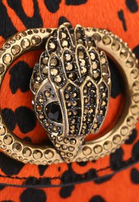 Spilla in metallo dorato e nero a forma di mano, impreziosita da strass neri e dorati, su uno sfondo con stampa leopardata arancione e nera.