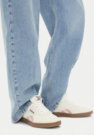 Zapatillas blancas con detalles en rosa y suela de goma marrón, combinadas con vaqueros de mezclilla azul claro de corte holgado. Los zapatos tienen diseño con cordones y logo.