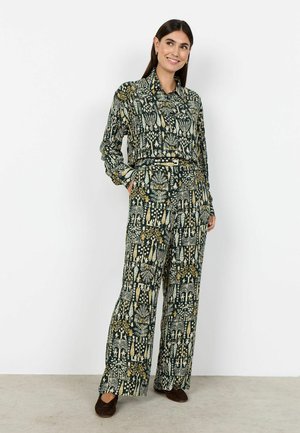 Femme portant une chemise boutonnée verte à motifs assortis et un pantalon à jambes larges, debout avec les mains dans les poches et souriante sur fond blanc.