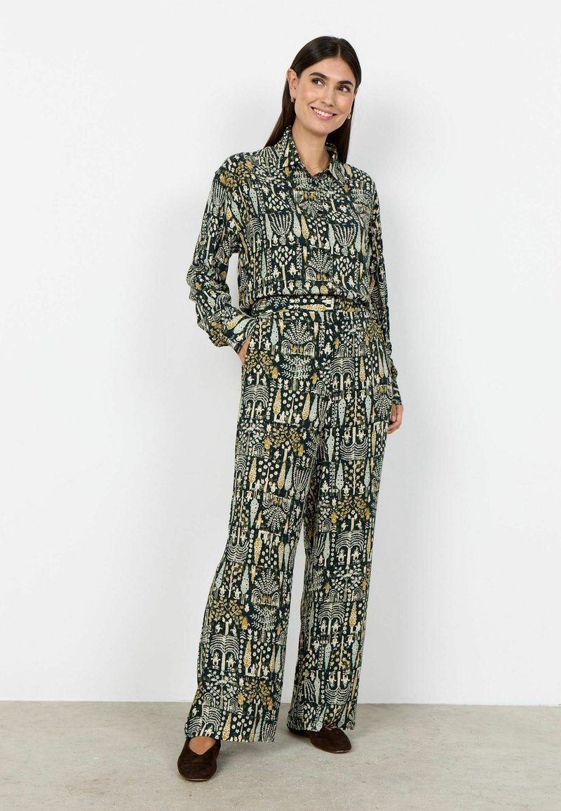 Femme portant une chemise boutonnée verte à motifs assortis et un pantalon à jambes larges, debout avec les mains dans les poches et souriante sur fond blanc.