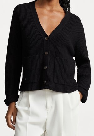 Žena v černém žebrovaném kardiganu na knoflíky se dvěma předními kapsami a bílých plisovaných kalhotách, ruce v kapsách.