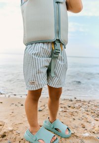 Gaiši zils zipa vesti ar ribveida tekstūru, kopā ar baltu un gaiši zilu svītrainu šortu pāri. Kājas tirkīza krāsas slip-on sandales.