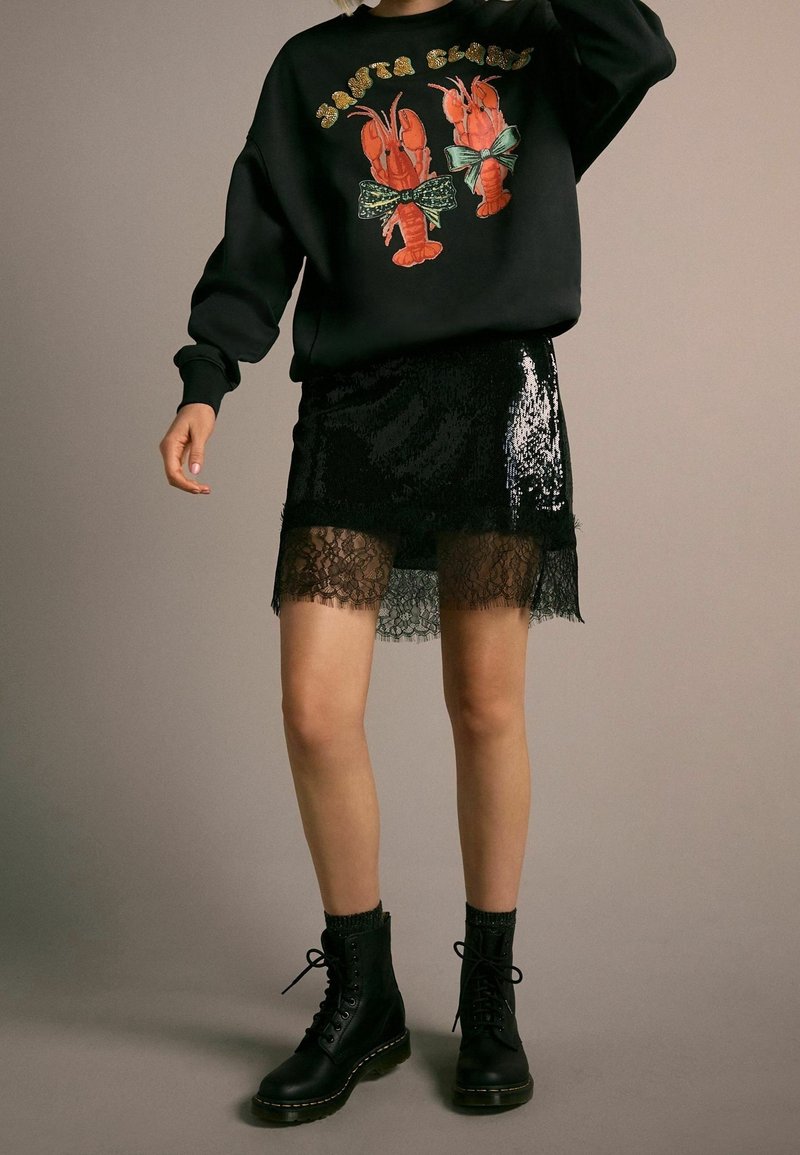 Personne portant un sweat-shirt noir avec deux homards rouges et une mini-jupe noire à sequins avec bordure en dentelle, associée à des bottes noires à lacets.
