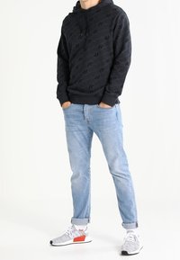 Osoba oblečená v černé mikině Adidas, světle modrých vykasaných džínách a bílých teniskách s oranžovými akcenty, stojící s rukama v kapsách.