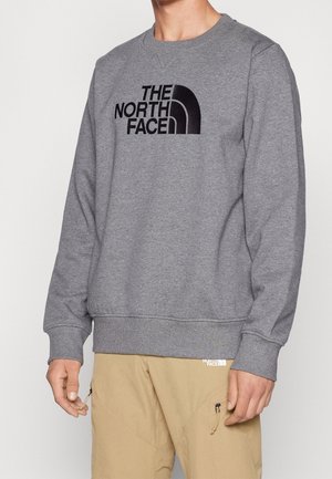 Mand iført grå langærmet sweatshirt med "The North Face" logo og beige bukser med lynlåslommer, stående mod en ensfarvet baggrund.