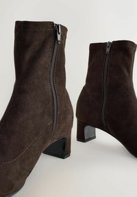 Een paar donkerbruine suède enkellaarzen met ritssluitingen aan de zijkant en lage blokhakjes op een effen lichte achtergrond.