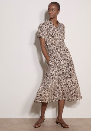 Kobieta w brązowo-białej sukience midi w kropki, z krótkimi rękawami i kieszeniami, stoi z rękami w kieszeniach i patrzy w prawo.