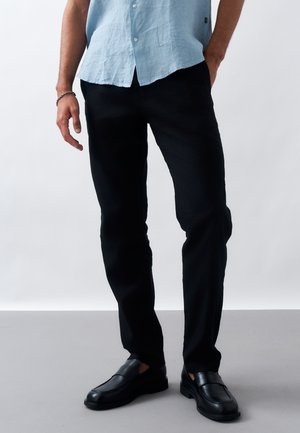 Άνδρας που φοράει μαύρο παντελόνι, μαύρα μοκασίνια και ανοιχτό μπλε κοντομάνικο πουκάμισο με το ένα χέρι στην τσέπη.