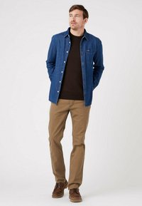 Camicia in denim blu con chiusura a bottoni e taschino sul petto, sovrapposta a un maglione marrone scuro. Pantaloni marrone chiaro e scarpe marroni completano l'outfit.