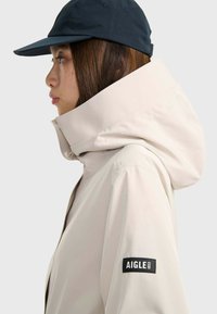 Veste imperméable beige avec un col haut et une grande capuche, arborant un patch logo Aigle noir sur la manche. Portée avec une casquette sombre.