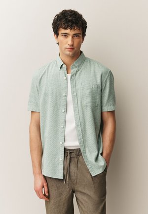 Ung mand står mod en neutral baggrund iført en grøn ternet kortærmet skjorte over en hvid tanktop og brune bukser med snor i taljen.