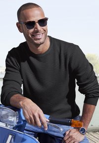 Schwarzer strukturierter Pullover mit gerippten Bündchen, getragen von einer Person, die auf einem blauen Roller sitzt. Auffällige Accessoires sind braune Sonnenbrillen und eine Uhr.