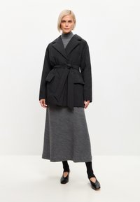 Abrigo negro con cinturón, con cuello de muesca y bolsillos, combinado con un vestido largo gris, medias negras y zapatos negros.